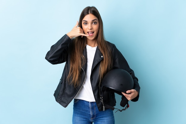 Giovane donna caucasica che tiene un casco del motociclo isolato sulla parete blu che fa gesto del telefono. Richiamami segno