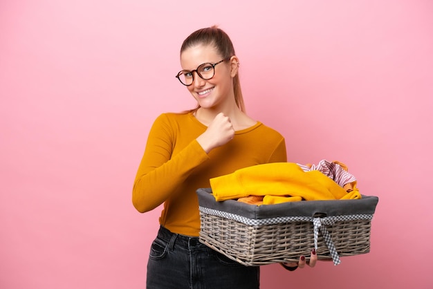 Giovane donna caucasica che tiene un cesto di vestiti isolato su sfondo rosa che celebra una vittoria