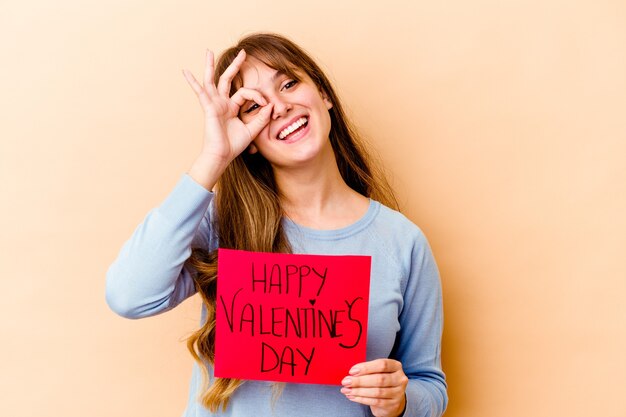 Giovane donna caucasica che tiene un felice giorno di biglietti di S. Valentino isolato eccitato mantenendo il gesto giusto sull'occhio.