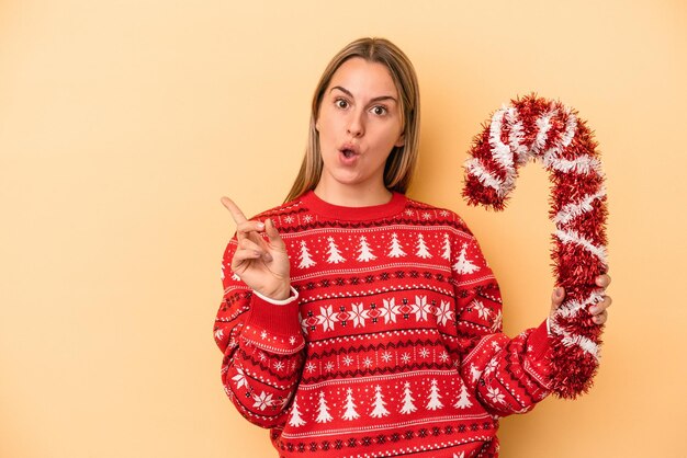 Giovane donna caucasica che tiene un grande bastone di natale isolato su sfondo giallo che punta al lato