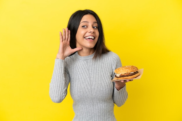 Giovane donna caucasica che tiene un hamburger isolato su sfondo giallo ascoltando qualcosa mettendo la mano sull'orecchio