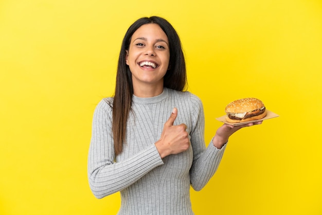 Giovane donna caucasica che tiene un hamburger isolato su sfondo giallo che dà un gesto di pollice in alto