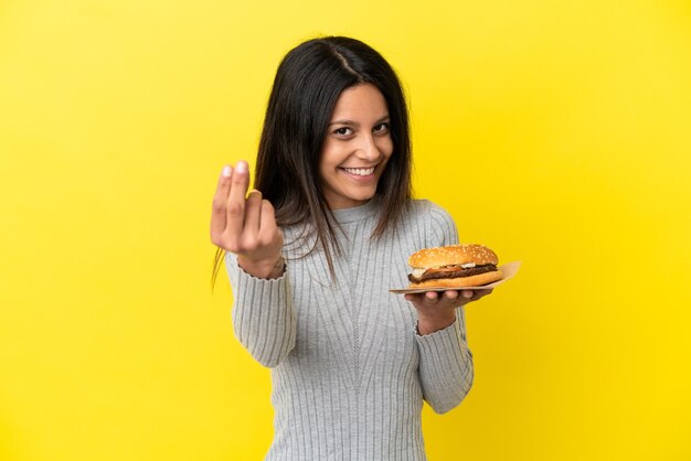 Giovane donna caucasica che tiene un hamburger isolato su sfondo giallo che fa gesto di denaro