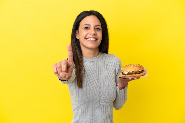 Giovane donna caucasica che tiene un hamburger isolato su sfondo giallo che mostra e solleva un dito