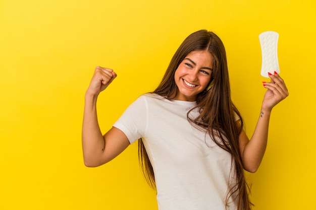 Giovane donna caucasica che tiene un impacco isolato su sfondo giallo alzando il pugno dopo una vittoria, concetto vincitore.