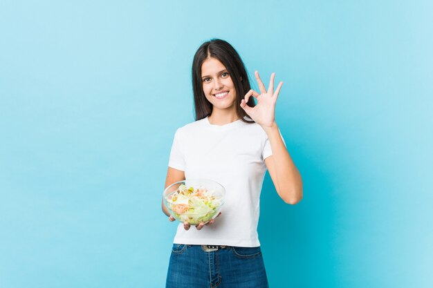 Giovane donna caucasica che tiene un'insalata allegra e sicura che mostra gesto giusto.