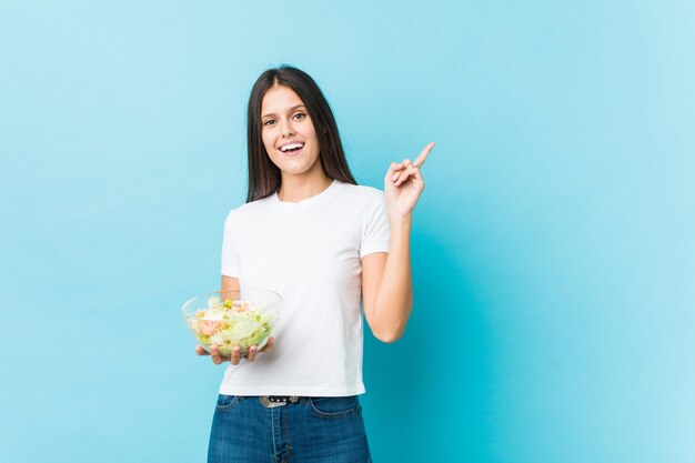 Giovane donna caucasica che tiene un'insalata che sorride allegramente indicando con l'indice di distanza.