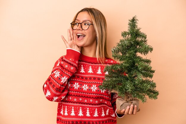 Giovane donna caucasica che tiene un piccolo albero di natale isolato su sfondo beige gridando e tenendo il palmo vicino alla bocca aperta.