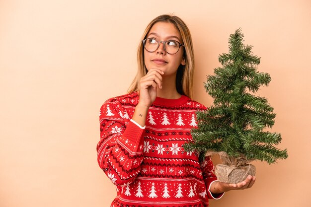 Giovane donna caucasica che tiene un piccolo albero di natale isolato su sfondo beige guardando di traverso con espressione dubbiosa e scettica.