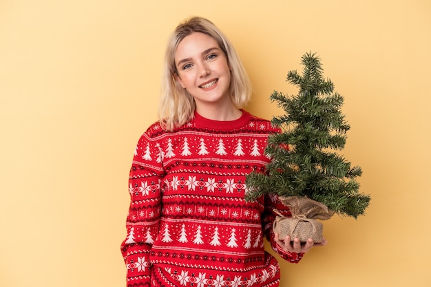 Giovane donna caucasica che tiene un piccolo albero di natale isolato su sfondo giallo felice, sorridente e allegro.