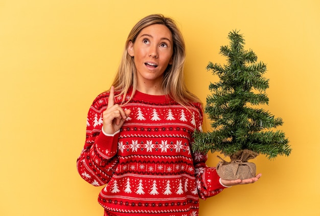 Giovane donna caucasica che tiene un piccolo albero di natale isolato su sfondo giallo rivolto verso l'alto con la bocca aperta.