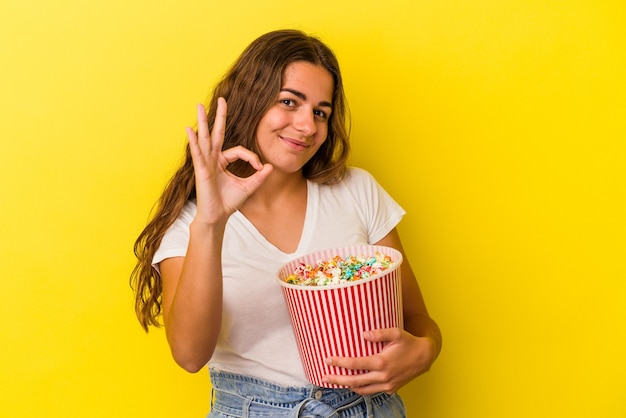 Giovane donna caucasica che tiene un popcorn isolato su sfondo giallo allegro e fiducioso che mostra gesto ok.