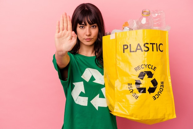 Giovane donna caucasica che tiene un sacchetto di plastica riciclato isolato su sfondo rosa in piedi con la mano tesa che mostra il segnale di stop, impedendoti.