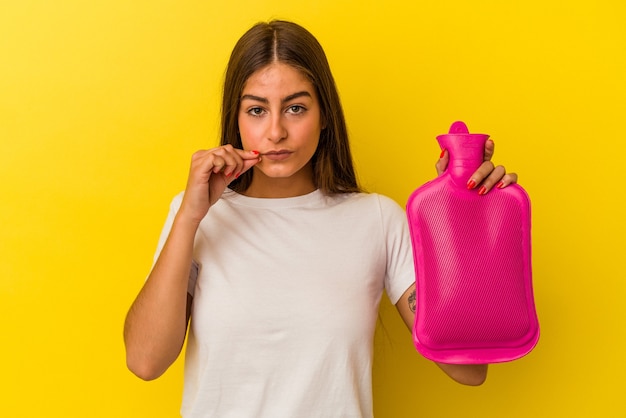 Giovane donna caucasica che tiene una bottiglia d'acqua calda isolata su sfondo giallo con le dita sulle labbra mantenendo un segreto.