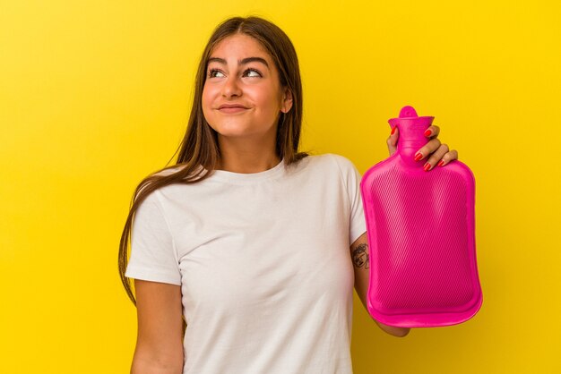 Giovane donna caucasica che tiene una bottiglia d'acqua calda isolata sul muro giallo sognando di raggiungere obiettivi e scopi