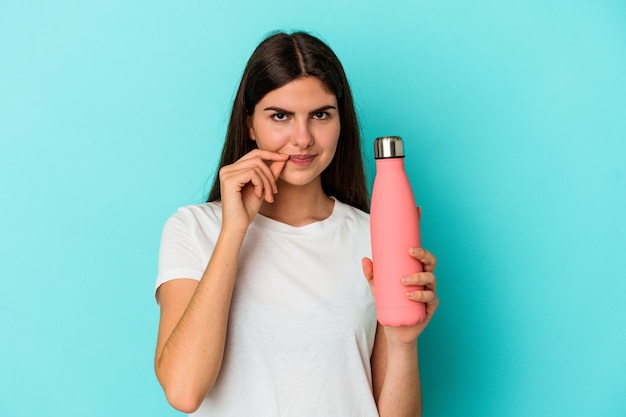 Giovane donna caucasica che tiene una bottiglia d'acqua isolata su sfondo blu con le dita sulle labbra mantenendo un segreto.