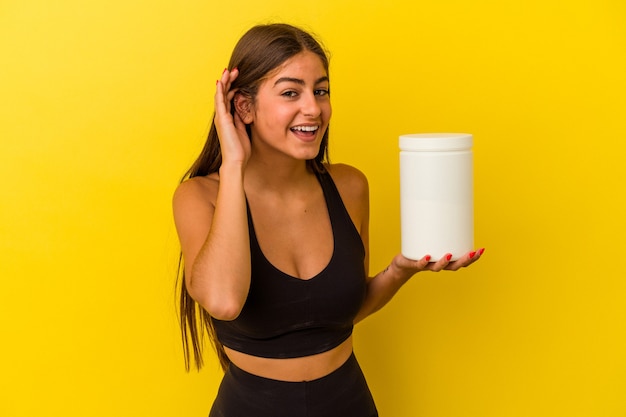 Giovane donna caucasica che tiene una bottiglia di proteine isolata su sfondo giallo cercando di ascoltare un pettegolezzo.