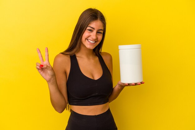 Giovane donna caucasica che tiene una bottiglia di proteine isolata su sfondo giallo gioiosa e spensierata che mostra un simbolo di pace con le dita.