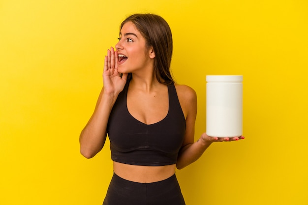 Giovane donna caucasica che tiene una bottiglia di proteine isolata su sfondo giallo gridando e tenendo il palmo vicino alla bocca aperta.