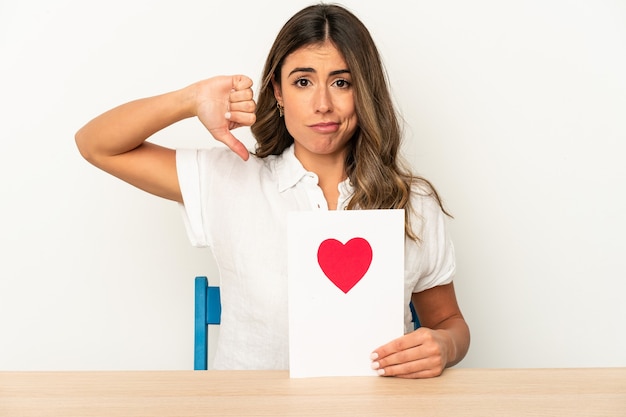 Giovane donna caucasica che tiene una carta di San Valentino isolata che mostra un gesto di avversione, pollice in giù. Concetto di disaccordo.