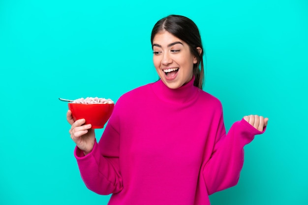 Giovane donna caucasica che tiene una ciotola di cereali isolata su sfondo blu che celebra una vittoria