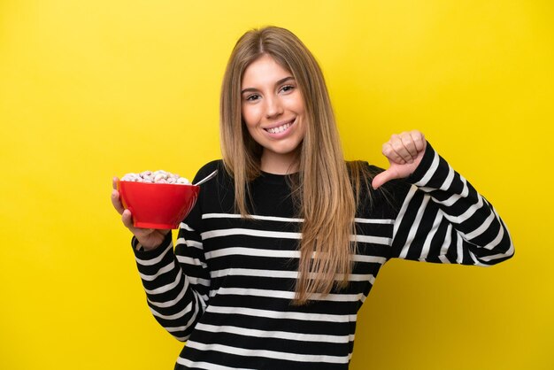 Giovane donna caucasica che tiene una ciotola di cereali isolata su sfondo giallo orgogliosa e soddisfatta