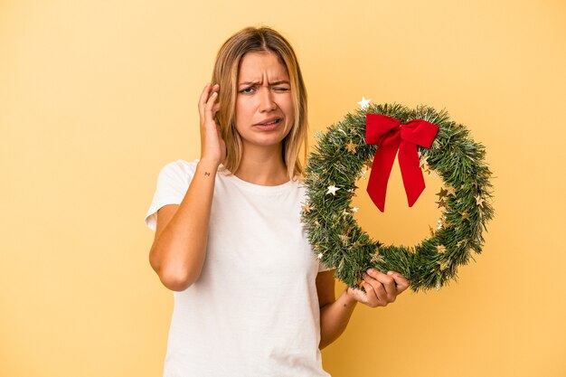 Giovane donna caucasica che tiene una corona di Natale isolata su sfondo giallo che copre le orecchie con le mani.