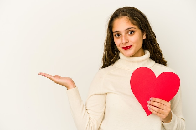 Giovane donna caucasica che tiene una forma di San Valentino del cuore isolata che mostra uno spazio della copia su una palma e che tiene un'altra mano sulla vita.