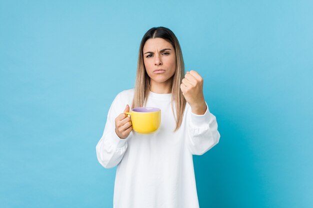 Giovane donna caucasica che tiene una tazza di caffè che mostra pugno alla macchina fotografica, espressione facciale aggressiva.