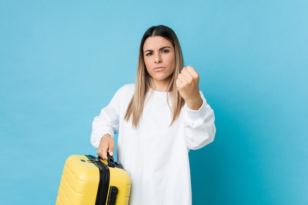 Giovane donna caucasica che tiene una valigia che mostra pugno alla macchina fotografica, espressione facciale aggressiva.