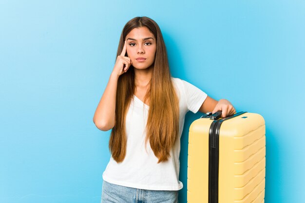 Giovane donna caucasica che tiene una valigia di viaggio che indica tempio con il dito, pensando, incentrato su un compito.