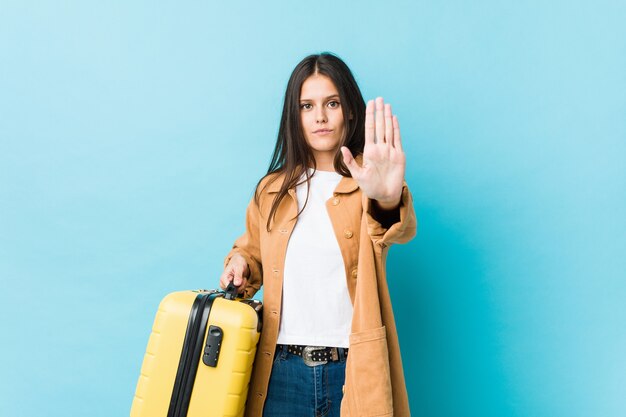 Giovane donna caucasica che tiene una valigia in piedi con la mano tesa che mostra il segnale di stop, impedendoti.