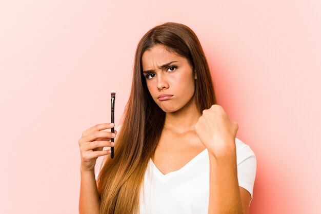 Giovane donna caucasica che tiene uno skinbrush che mostra pugno alla macchina fotografica, espressione facciale aggressiva.