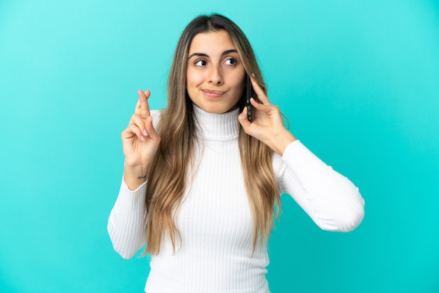 Giovane donna caucasica che utilizza il telefono cellulare isolato su sfondo blu con le dita incrociate e augurando il meglio