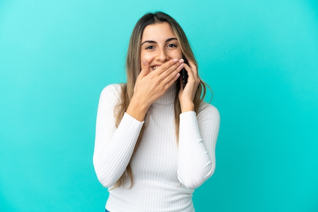 Giovane donna caucasica che utilizza il telefono cellulare isolato su sfondo blu felice e sorridente che copre la bocca con la mano