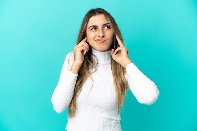 Giovane donna caucasica che utilizza il telefono cellulare isolato su sfondo blu frustrato e che copre le orecchie