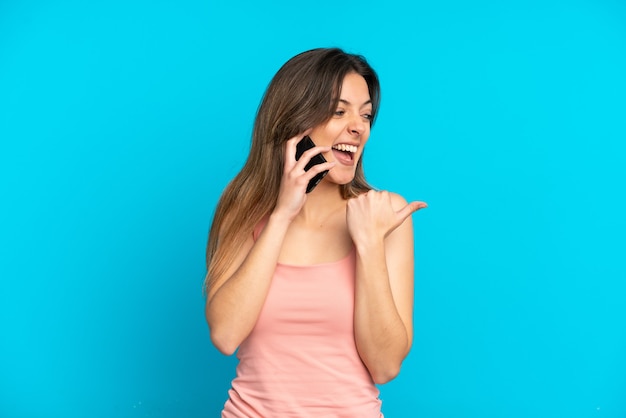 Giovane donna caucasica che utilizza il telefono cellulare isolato su sfondo blu rivolto verso il lato per presentare un prodotto