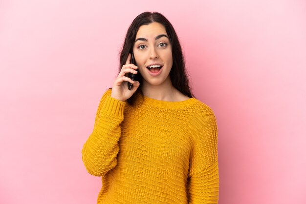 Giovane donna caucasica che utilizza il telefono cellulare isolato su sfondo rosa con espressione facciale a sorpresa