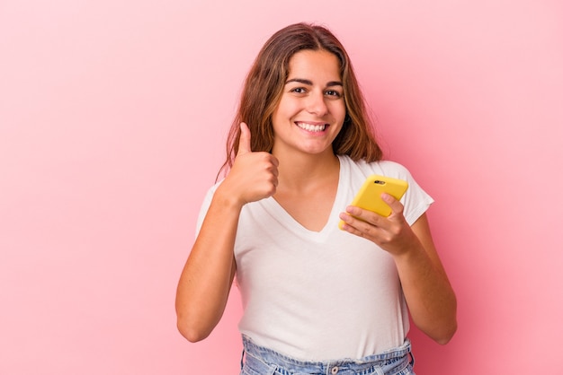 Giovane donna caucasica che utilizza il telefono cellulare isolato su sfondo rosa sorridente e alzando il pollice