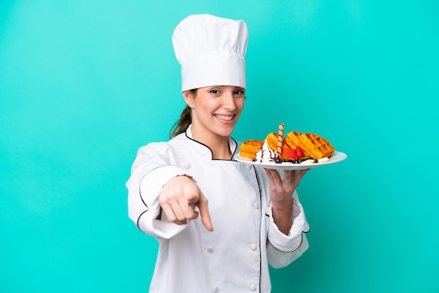 Giovane donna caucasica chef che tiene cialde isolate su sfondo blu punta il dito contro di te con un'espressione sicura