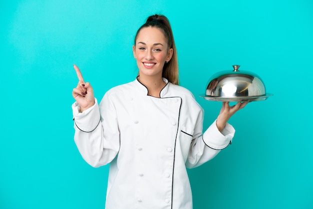 Giovane donna caucasica chef con vassoio isolato su sfondo blu che mostra e alza un dito in segno di meglio