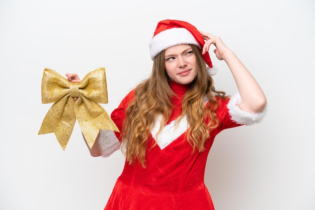 Giovane donna caucasica con abito di Natale che tiene fiocco di Natale isolato su sfondo bianco con dubbi e con espressione facciale confusa