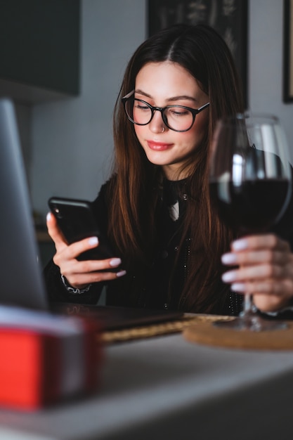 Giovane donna caucasica con capelli castani che riposa a casa, bere vino e utilizzare smartphone e laptop