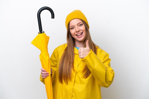 Giovane donna caucasica con cappotto antipioggia e ombrello isolati su sfondo bianco con i pollici in su perché è successo qualcosa di buono