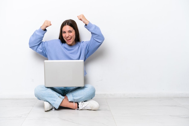 Giovane donna caucasica con laptop seduto sul pavimento isolato su sfondo bianco che celebra una vittoria in posizione di vincitore