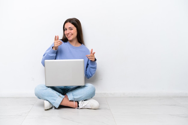 Giovane donna caucasica con laptop seduto sul pavimento isolato su sfondo bianco che punta in avanti e sorridente