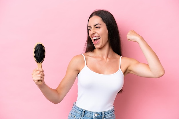 Giovane donna caucasica con pettine per capelli isolato su sfondo rosa che celebra una vittoria