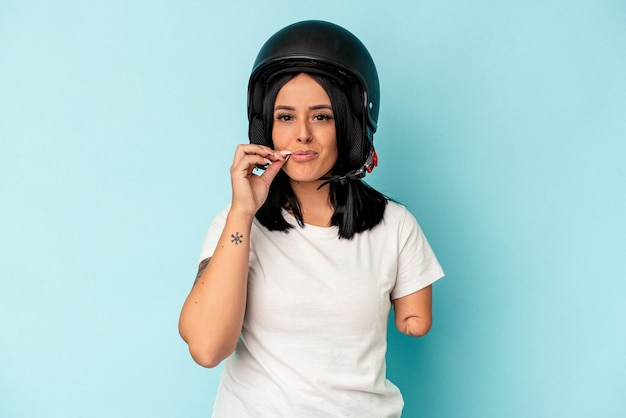 Giovane donna caucasica con un braccio che indossa un casco da motociclista isolato o sfondo blu con le dita sulle labbra mantenendo un segreto.