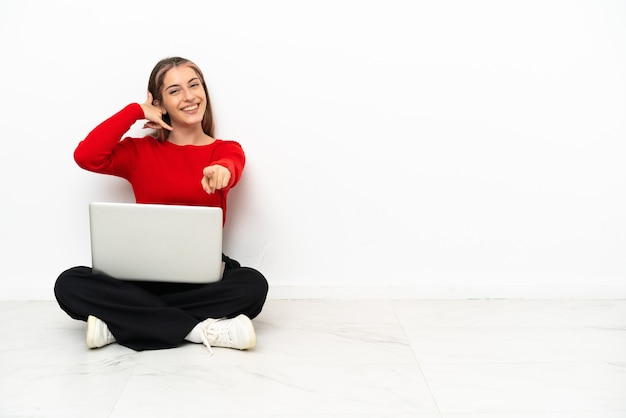 Giovane donna caucasica con un computer portatile che si siede sul pavimento che fa il gesto del telefono
