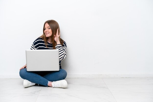 Giovane donna caucasica con un computer portatile seduto sul pavimento isolato su sfondo bianco ascoltando qualcosa mettendo la mano sull'orecchio